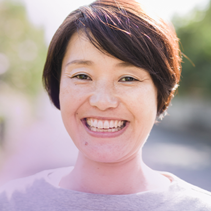 A close-up of a smiling woman with short, dark hair standing outside on a sunny day. The woman is dressed casually and her broad smile conveys a sense of happiness and positivity.