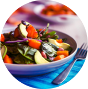 Fresh salad in a white bowl. The salad includes vibrant ingredients such as leafy greens, avocado slices, and bright orange vegetables, possibly sweet potatoes or carrots. 
