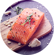 A raw salmon fillet placed on a wooden cutting board. The salmon is garnished with sprigs of herbs and black pepper, surrounded by garlic cloves and sprinkled with seasoning, suggesting preparation for cooking.