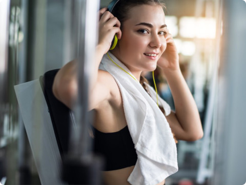 Een jonge vrouw in een zwarte sportbeha zit op een fitnessapparaat in de sportschool en lacht terwijl ze haar koptelefoon opzet. Een witte handdoek is om haar nek gedrapeerd en ze lijkt een pauze te nemen van de training. Op de achtergrond staan fitnessapparaten met zonlicht door de ramen.