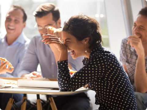 Een groep collega's deelt een luchtig moment terwijl ze pizza eten in een lichte kantoorruimte. Een vrouw op de voorgrond met een blouse met stippen lacht terwijl ze een stuk pizza vasthoudt. De anderen, gekleed in business casual kleding, glimlachen en praten rond een tafel.