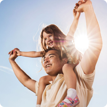Un père souriant porte sa jeune fille sur ses épaules, tandis qu'ils se tiennent par la main et lèvent les bras en l'air. Le soleil brille derrière eux, créant une lueur chaleureuse.