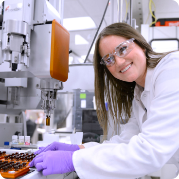 Une femme scientifique portant une blouse blanche, des lunettes de sécurité et des gants violets sourit alors qu'elle travaille dans un laboratoire. Elle manipule de petites fioles sous une machine robotisée, entourée d'équipements de laboratoire dans un environnement de haute technologie.