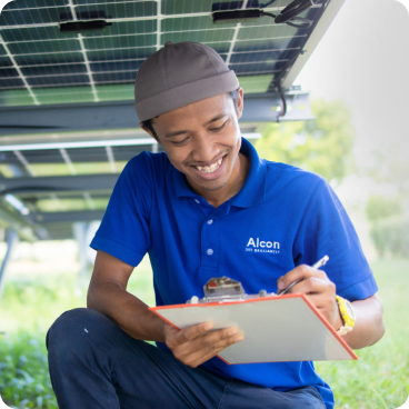 Un technicien portant un polo bleu et une casquette marron de la marque Alcon sourit tout en écrivant sur un bloc-notes. Il est accroupi en plein air sous une installation de panneaux solaires, entouré de verdure