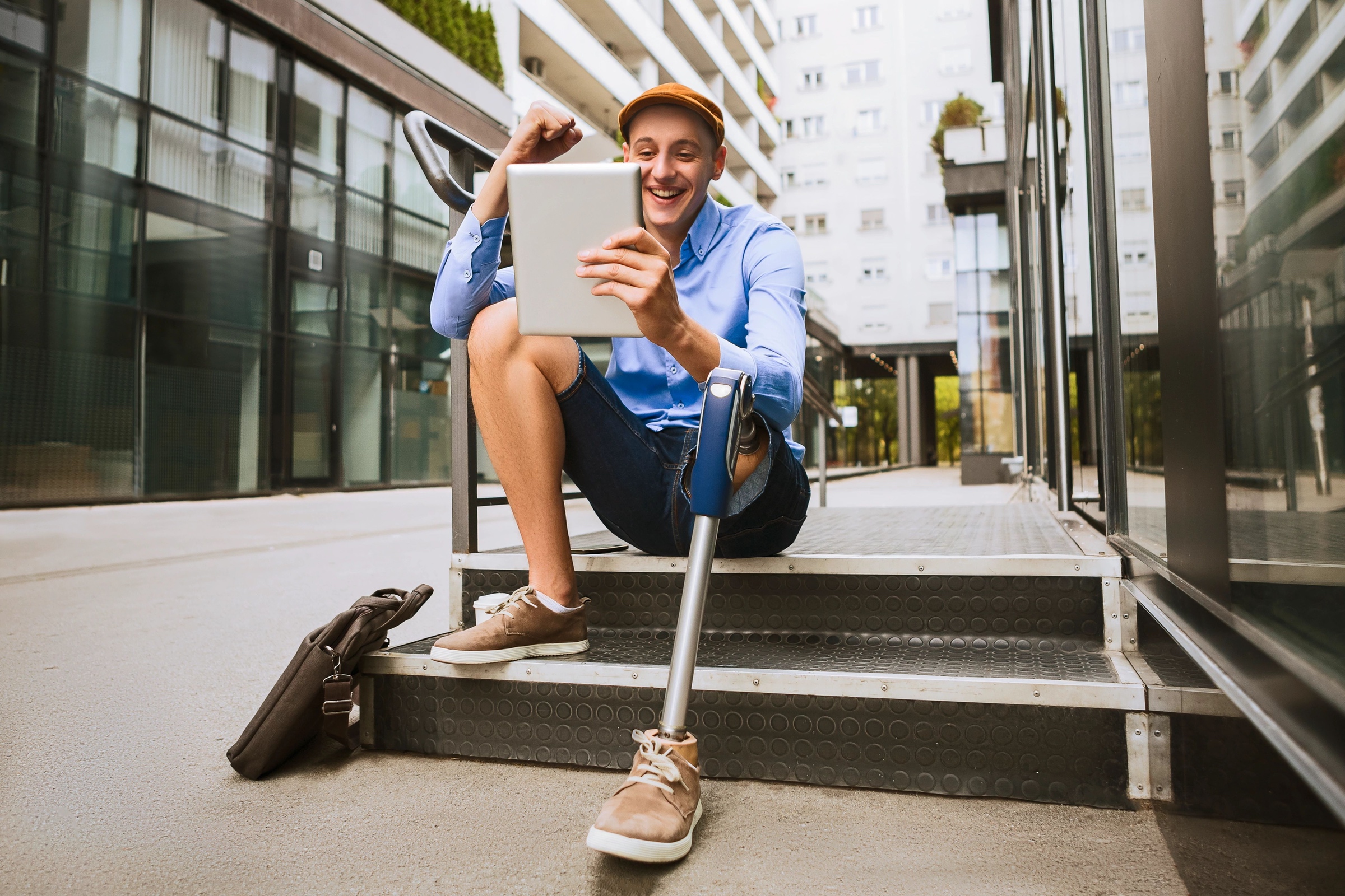 Young man with a prosthetic leg, sitting on stairs outside office building, smiling and looking at tablet